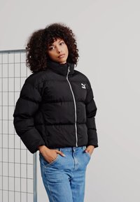 Svart boblejakke med høy krage, glidelås og hvit logo på brystet. Brukt over blå jeans i en minimalistisk innstilling.