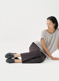 Svart skinn loafers med en blank finish, kombinert med en løs grå drapert topp og brede, mørkebrune bukser, på hvit bakgrunn.