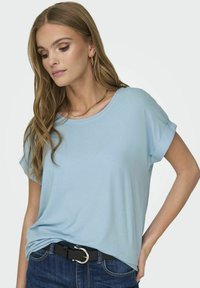 Lyseblå kortermet topp laget av mykt stoff, med løs passform og rullede ermer, sammen med mørkeblå jeans og et svart belte.