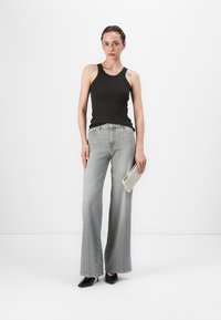 Jeans grii evazați, top negru ajustat și tocuri negre elegante. Persoana ține o poșetă albă texturată. Fundal minimalist.