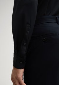 Maglietta nera a maniche lunghe con polsini a bottone e un logo testurizzato discreto sulla manica. Vestibilità aderente con tessuto morbido e design elegante.