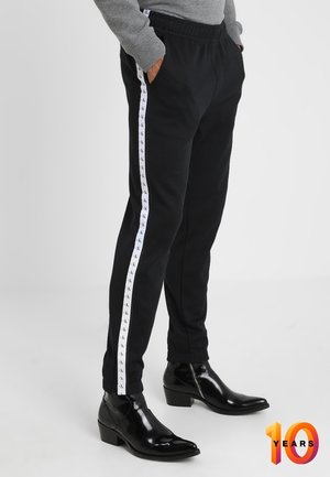 Persoon draagt zwarte broek met witte logostreep langs de zijkanten, grijze trui en zwarte glanzende leren enkellaarsjes.