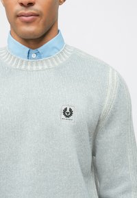 Ανοιχτό γκρι πλεκτό πουλόβερ με στρογγυλή λαιμόκοψη, ραβδωτές λεπτομέρειες και μια μικρή επιγραφή λογοτύπου Belstaff στο στήθος, σε στρώσεις πάνω από ένα μπλε πουκάμισο με γιακά.