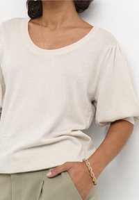 Kobieta w beżowej, krótkim rękawem dzianinowej bluzce, jasnoszarych spodniach, złotej skręconej bransoletce oraz delikatnym złotym łańcuszku, z ręką w kieszeni.