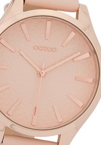 Round watch with a rose gold metal case, textured light pink dial, and rose gold hour markers. Smooth strap in light pink material.