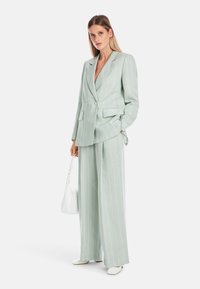 Hellgrüner Blazer mit Streifenmuster und weiten Hosen. Der Blazer hat doppelte Knöpfe und Taschen. Das Modell trägt eine weiße Handtasche.
