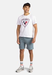 Camiseta blanca con logo de Rossignol en rojo y negro, combinada con pantalones cortos azules. El modelo lleva zapatillas deportivas grises y calcetas de rayas múltiples. Diseño casual.