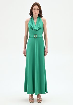 Donna che indossa un abito verde in raso senza maniche con scollo a halter drappeggiato, vita cintata e sandali dorati con tacco alto e cinturini.