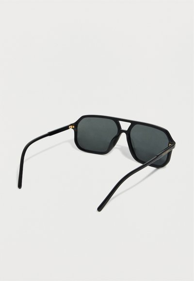 Lunettes de soleil aviateur noires avec des verres foncés et un pont nasal fin, vues en angle arrière sur un fond clair.