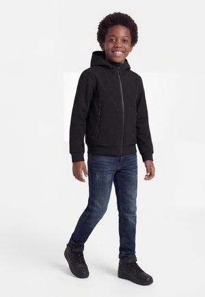 Χαμογελαστό αγόρι που φοράει μαύρο μπουφάν με κουκούλα, μπλε τζιν και μαύρα ψηλά αθλητικά παπούτσια, όρθιο με το ένα πόδι ελαφρώς μπροστά.