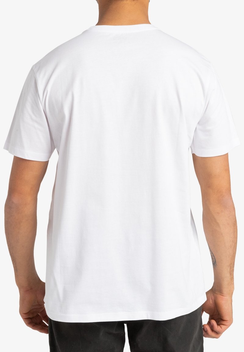 Homme portant un t-shirt blanc uni à manches courtes vu de dos, pantalon noir, debout sur un fond blanc.