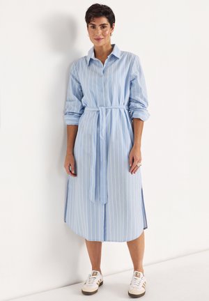 Frau steht vor einer weißen Wand und trägt ein hellblaues, langärmliges, tailliertes, gestreiftes Hemdblusenkleid und weiße Turnschuhe.