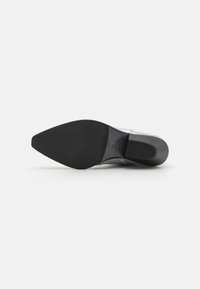 Suela de goma negra de un zapato de tacón alto con puntera afilada. Se pueden ver detalles en plata en el tacón y en la parte superior. Textura suave en general.