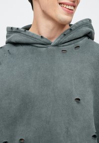 Sivá mikina s kapucňou s opotrebovanými dierami, viditeľnou textúrou látky a voľným strihom s kapucňou na šnúrky. Detailný záber na oblasť výstrihu a hrudníka.