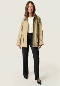 Veste légère beige avec un col marron, deux poches avant et une fermeture à glissière, assortie à un pantalon noir tailleur et des chaussures noires à bouts pointus.