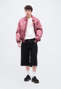Νεαρός άνδρας που φορά ροζ μπουφάν τύπου bomber, λευκό μπλουζάκι με τύπωμα, μαύρα φαρδιά σορτς, λευκές κάλτσες και μαύρα υποδήματα slip-on, στέκεται σε λευκό φόντο.