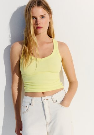 Jeune femme aux longs cheveux blonds portant un crop top jaune sans manches et un pantalon blanc taille haute, debout contre un mur blanc.