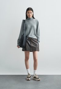 Grauer Langarm-Rollkragenpullover, brauner Minirock aus Leder mit sichtbaren Nähten, schwarze Schultertasche, weiße Socken und klobige Sneakers.