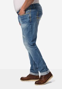 Jeans de denim azul claro con áreas desgastadas y detalles desgastados. Cuenta con cinco bolsillos y un dobladillo enrollado. Combinados con botas marrones con cordones.