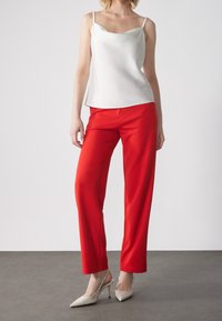 Camisola blanca de satén con tirantes finos combinada con pantalones rojos de talle alto; la modelo lleva zapatos slingback beige. Tela suave, diseño elegante.