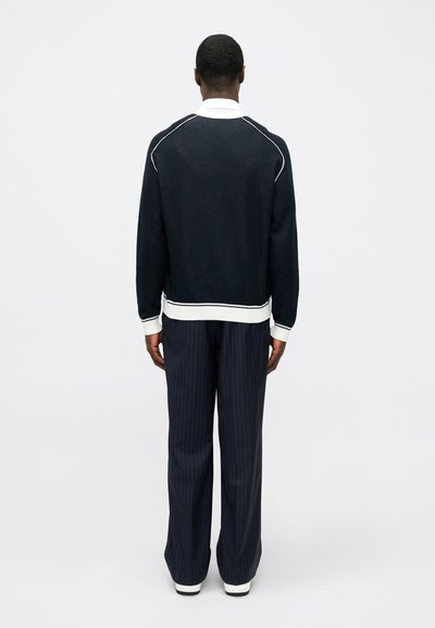 Άνδρας που στέκεται με την πλάτη, φορώντας μαύρη μπλούζα με μακριά μανίκια και λευκό γιακά και μανσέτες, σκούρες ρίγες παντελόνι και λευκά παπούτσια.