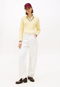 Κίτρινο πουλόβερ με πλέξη καλτσοδέτας και λαιμόκοψη σε σχήμα V, λευκό πουκάμισο με γιακά από κάτω, φαρδιά λευκά παντελόνια και καφέ μοκασίνια με διακοσμητικές φούντες. Μπορντό καπέλο.