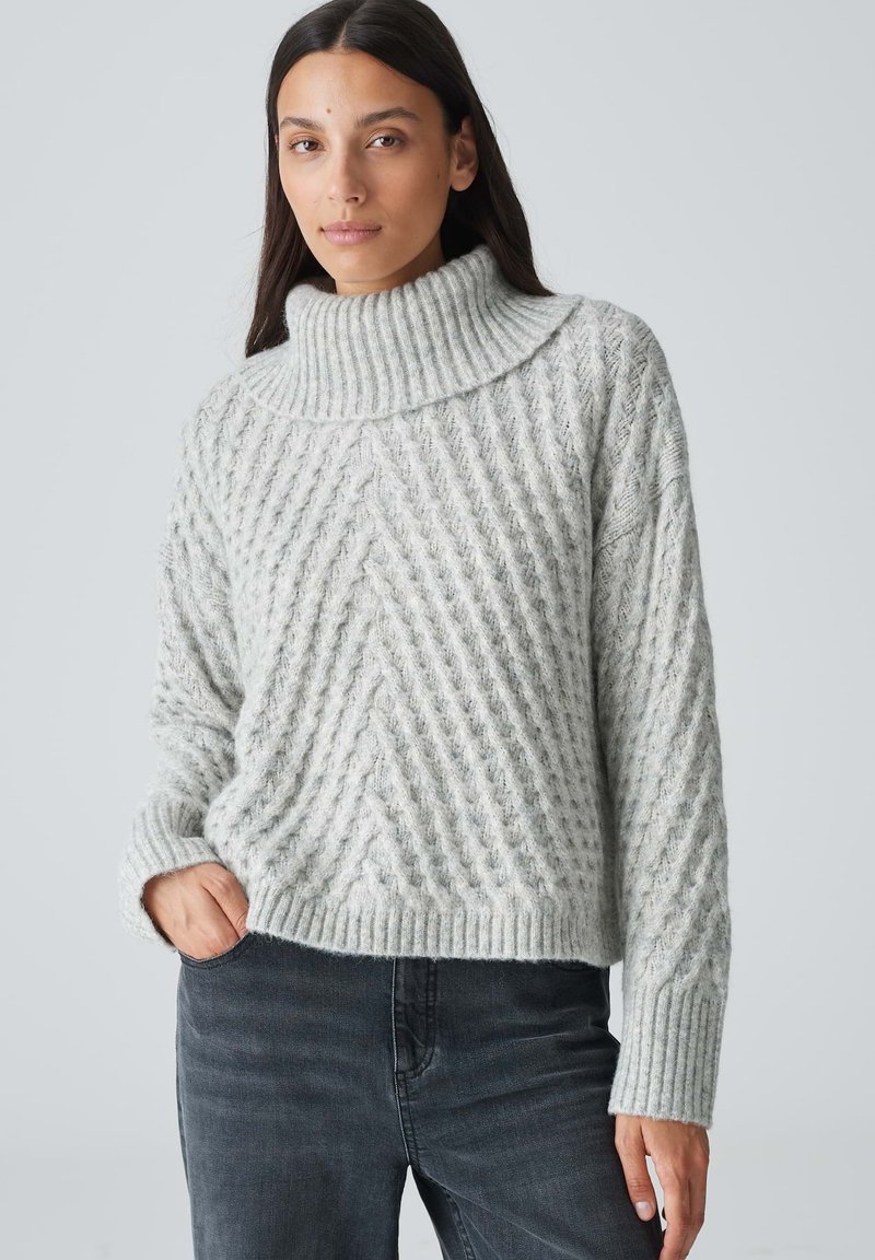 Grauer Strickpullover mit hohem Kragen, der ein strukturiertes Zickzackmuster und gerippte Bündchen aufweist. Getragen mit dunklen Jeans.
