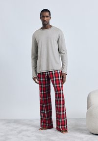 Pelēka garām piedurknēm krekls un sarkanas raibu pidžamas bikses no mīksta auduma, ar brīvu piegriezumu un redzamām šuvēm. Kājās basas uz gaiša paklāja.