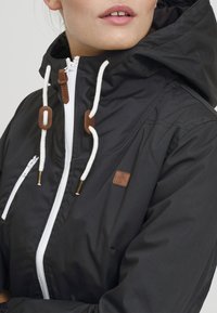 Svart hettejakke med glidelås, med hvite snorer, brune lærdetaljer og en liten logo på venstre brystområde.