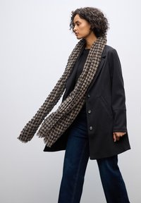 Cappotto di lana nera con grandi bottoni, abbinato a una sciarpa marrone e nera a motivo. La sciarpa presenta dettagli con frange e una trama testurizzata.