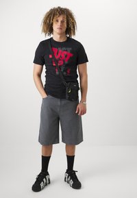 Svart t-skjorte med rød grafikk, grå denimshorts, svart crossover-veske, svarte sneakers med hvite detaljer, og svarte sokker. Rettstående posisjon.