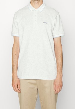 Mann trägt ein hellgraues Polohemd mit kurzen Ärmeln und dem "BOSS"-Logo auf der Brust sowie beige Hosen, steht vor weißem Hintergrund.