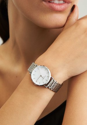 Montre-bracelet en maille argentée avec un cadran circulaire blanc, des index horaires minimalistes et un boîtier en acier inoxydable élégant.