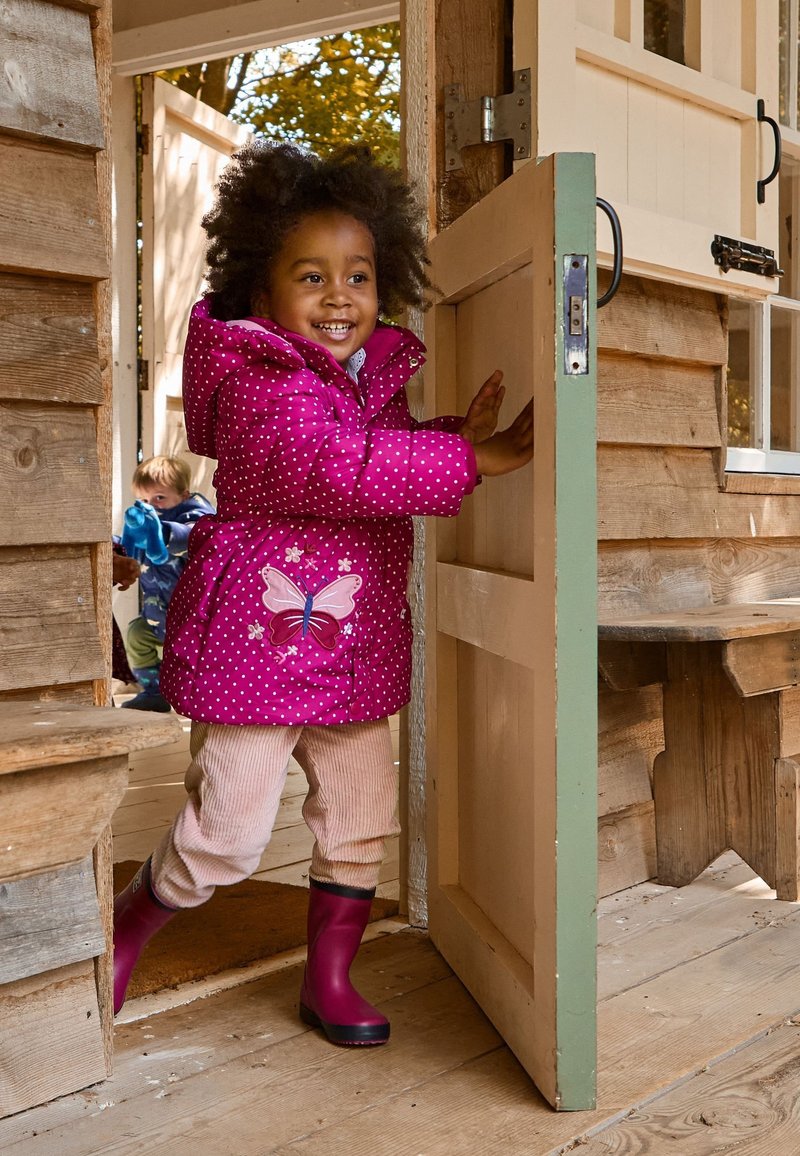 Roze gewatteerde jas met stippen en een vlinderpatroon, beige ribfluwelen broek en roze rubberen laarzen. Meisje staat bij een houten deur.