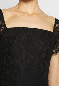 Abito in pizzo nero con scollatura quadrata e maniche corte in pizzo decorativo. Il tessuto presenta un motivo floreale texturizzato.