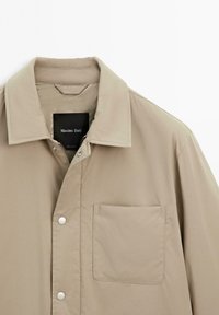 Giacca beige con chiusura a bottoni, colletto ampio e una tasca sul petto. Realizzata in tessuto liscio e leggero. Caratterizzata da bottoni a pressione in metallo.