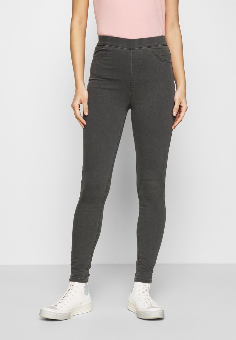 Femme portant un jean skinny gris foncé, un haut rose pâle et des baskets montantes blanches, se tenant devant un fond uni.