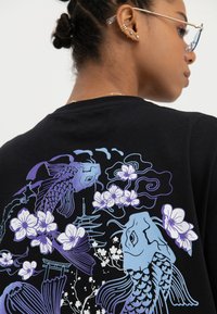 Frau trägt ein schwarzes Hemd mit blauem und lila Koi-Fisch, Kirschblüten und einem Pagoden-Design auf dem Rücken, von hinten gesehen.