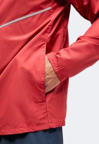 Main insérée dans la poche latérale d'une veste rouge avec bande réfléchissante, portée sur un pantalon bleu foncé.