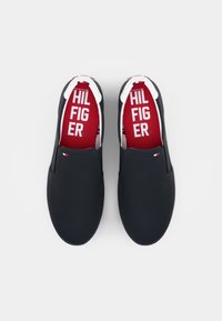 Pár námořnických slip-on bot s bílými podrážkami a červenými stélkami potištěnými bílým textem "HILFIGER", zobrazené z vrchu na bílém pozadí.