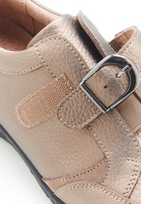 Scarpa in pelle bronzo con finitura testurizzata, dotata di una cinghia in tessuto e fibbia metallica. Forma arrotondata e dettagli di cucitura a contrasto.