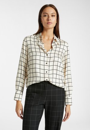 Blusa color crema con motivo a griglia nera, dotata di colletto, maniche lunghe e bottoni neri, abbinata a pantaloni a scacchi neri.