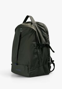 Mochila de tela verde con una textura suave, que cuenta con un bolsillo con cremallera en la parte frontal, correas ajustables y un acento de logo cerca de la parte superior.