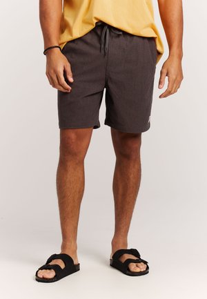 Man draagt donkergrijze, getextureerde shorts met trekkoord, gele shirt, zwarte sandalen met banden, staand tegen een effen lichte achtergrond.