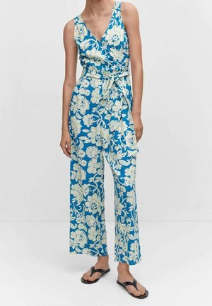 Femme portant une combinaison bleue sans manches avec un motif floral blanc et une ceinture à la taille, associée à des sandales noires.