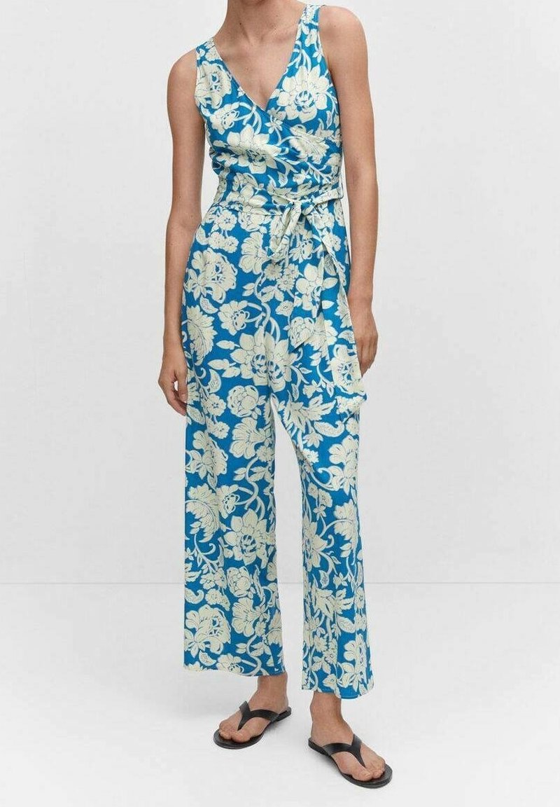 Femme portant une combinaison bleue sans manches avec un motif floral blanc et une ceinture à la taille, associée à des sandales noires.