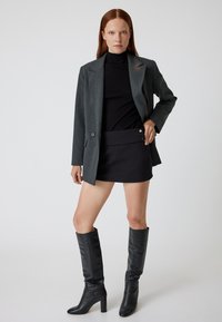 Blazer grigio su misura, dolcevita nero aderente, mini gonna nera con bottoni a pressione e stivali neri di pelle con tacco al ginocchio.