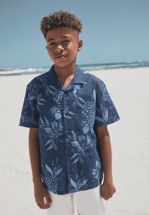 Junge mit lockigem Haar, der ein blaues, kurzärmeliges Hemd mit Blumenmuster und weiße Shorts trägt, steht an einem sandigen Strand mit Meereswellen im Hintergrund.