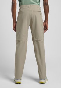 Beige Zip-Off-Hosen aus glattem, leichtem Stoff; elastischer Taillenbund; und eine Reißverschlusstasche auf der Rückseite. Abgerundet mit grauen Schuhen.