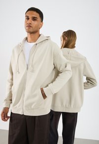 Beige mikina na zip s kapucí, předními kapsami a žebrovanými manžetami. Model stojí čelem vpřed s další osobou v pozadí.